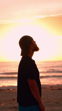 Silhouette of a pensive bearded man standing on the beach watching the sunset, feeling freedom and tranquility while contemplating the serene ocean waves and golden hour light. Christian man praying. 
