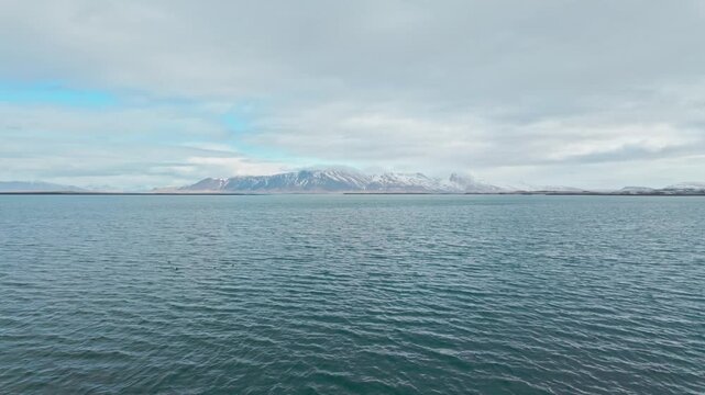 A landscape view of Mt Esja in Iceland behind a foreground of a vast and open bay of blue water under a bright cloudy sky.