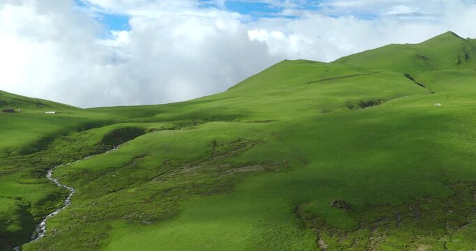 Badimalika region, Bajura, Nepal, aerial view over lush green alpine pastures, stream beds, and rolling hills in Triveni Patan grassland under blue skies and white cloud formations.