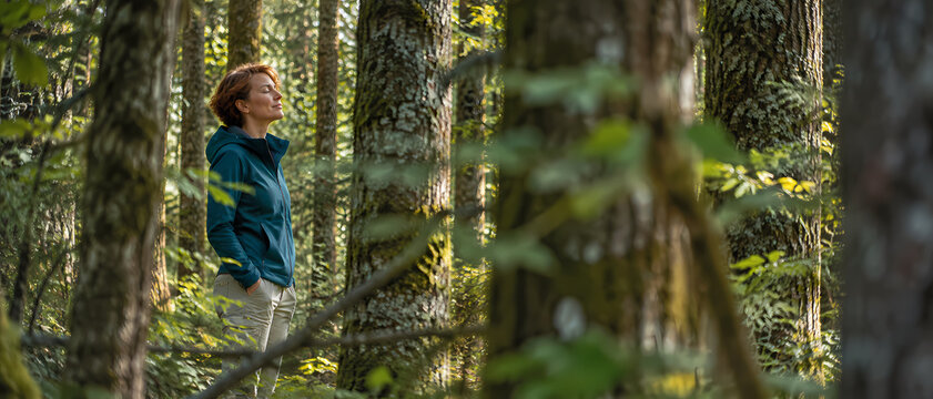 Relaxed woman stands among tall trees in a lush green forest, breathing fresh air and enjoying a quiet mindful moment while soft sunlight filters through branches and woodland foliage around her