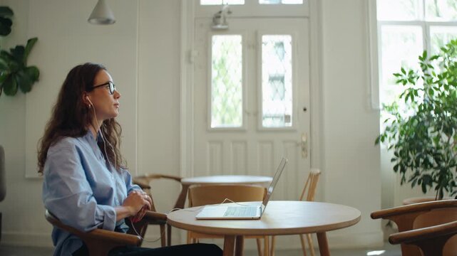 Woman joins a video call on her laptop in a bright cafe workspace, wearing earphones and gesturing as she speaks. The scene shows remote work, online meeting, freelance communication, and flexible wor