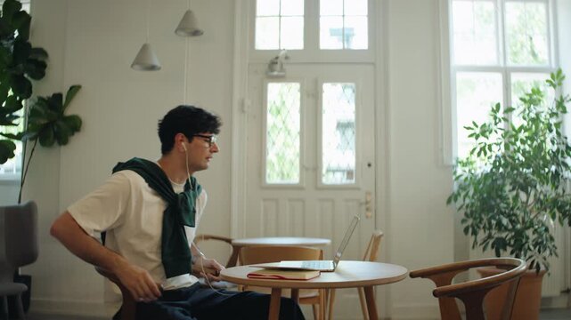 Man joins a laptop video call in a cafe workspace, speaking through earphones for remote work, online meeting tasks, freelance communication, and business discussion.