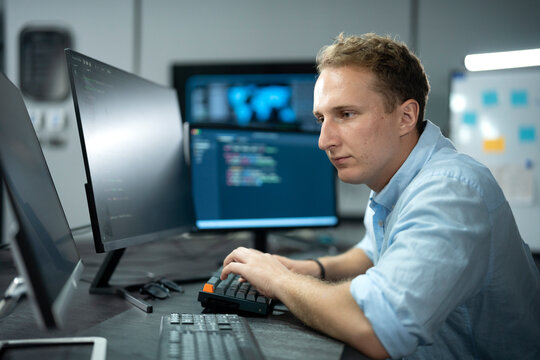 Male software engineer writing program code on multiple desktop screens in a modern office.