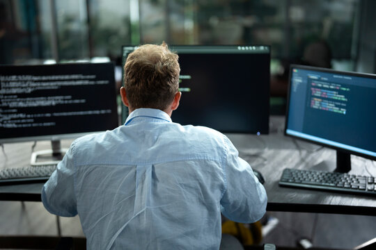 Rear view of male software engineer writing program code on multiple desktop screens in a modern office.
