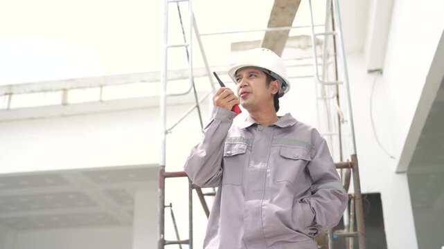 Male construction worker communicates by walkie talkie while looking around unfinished building and coordinating work activities with team at job site.