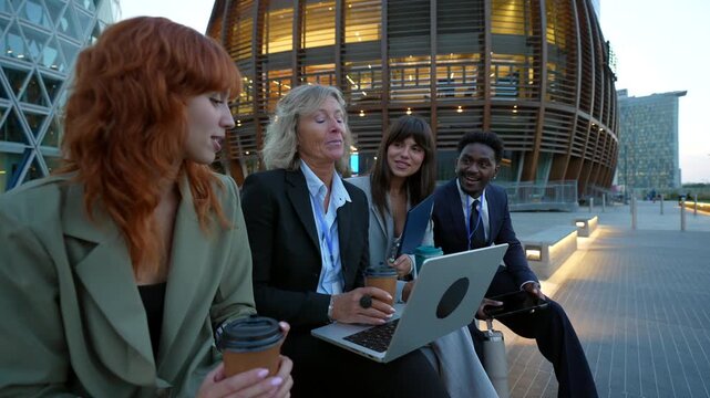Multiracial group of colleagues discussing a project on a laptop while sitting outside their modern office building, drinking coffee together and sharing ideas during a productive work break