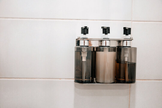 Three clear bottles in a metal holder dispensing shampoo, conditioner, and liquid soap on white bathroom tiles