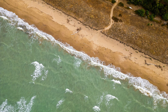A top-down aerial view highlights gentle white waves crashing along a wild, golden sandy beach in Molise, Italy. The contrast between the greenish turquoise sea and the natural coastal dunes creates a