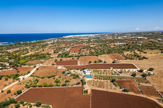 A high aerial view captures the rural Puglia countryside near Polignano a Mare, showcasing distinct red soil fields, scattered olive trees, and stone walls stretching towards the deep blue Adriatic