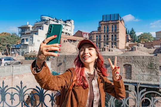Young woman traveling Abanotubani, Tbilisi taking a selfie on her smartphone, smiling and showing a peace sign on a sunny day