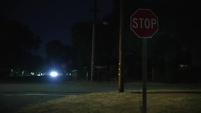 Car passes STOP sign at dark residential intersection late at night as bright headlights illuminate the road among trees, utility poles and deep shadows