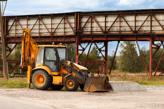 Yellow backhoe loader excavator parked under a massive weathered industrial steel conveyor gallery infrastructure structure at a mining quarry daylight horizontal view
