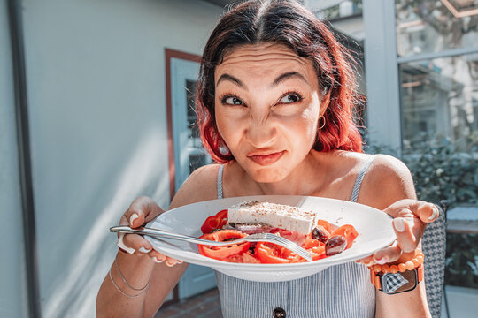 Playful reaction shot for food blogs and restaurant: woman making funny face while holding Greek salad plate with feta. Dining concept.
