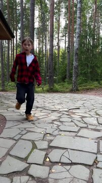 A vertical video showing a cute teenage boy of European appearance wearing a red checkered shirt and dark trousers walking along a stone path and then joyfully running.