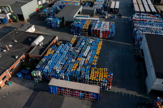 Aerial view of a large industrial facility featuring rows of stacked colorful chemical barrels, storage containers, and warehouse buildings in Roeselare, Flanders, Belgium.