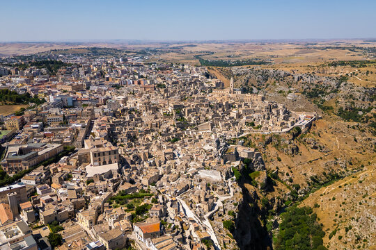 A high aerial panorama of Matera, Italy, showing the famous ancient Sassi quarter built along the edge of a dramatic canyon. The dense stone architecture, cathedral tower, and surrounding rugged