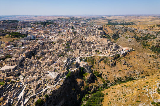 A high aerial panorama of Matera, Italy, showing the famous ancient Sassi quarter built along the edge of a dramatic canyon. The dense stone architecture, cathedral tower, and surrounding rugged