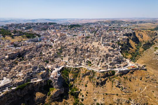 A high aerial panorama of Matera, Italy, showing the famous ancient Sassi quarter built along the edge of a dramatic canyon. The dense stone architecture, cathedral tower, and surrounding rugged