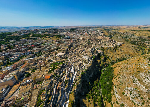 A high aerial panorama of Matera, Italy, showing the famous ancient Sassi quarter built along the edge of a dramatic canyon. The dense stone architecture, cathedral tower, and surrounding rugged