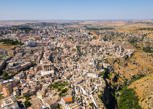 A high aerial panorama of Matera, Italy, showing the famous ancient Sassi quarter built along the edge of a dramatic canyon. The dense stone architecture, cathedral tower, and surrounding rugged