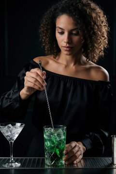 Attractive young woman in black off shoulder blouse stirs green cocktail with long metal spoon. Female bartender prepares refreshing alcoholic drink in glass at dark night club. Professional