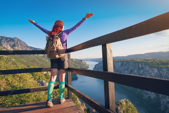 Arms-wide silhouette of a female hiker centered on a wooden overlook platform, framing a vivid Danube river Iron Gates canyon below. Adventure concept.
