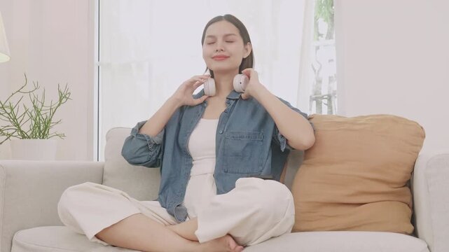 Happy Asian woman relaxing on sofa and listening to music with headphones, enjoying favorite song at home.
