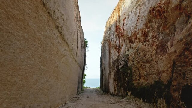 Narrow dirt path winding between towering limestone cliffs, revealing glimpse of the ocean and bright sky at the end, leading to Green Bowl Beach on the Bukit Peninsula, Bali island. Low drone flight