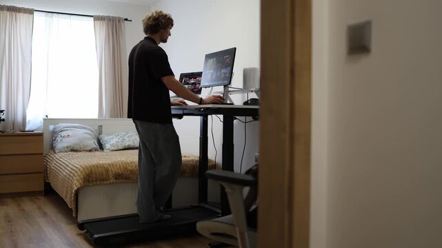 Young man works standing at an adjustable desk while browsing online content on dual screens choosing an upright workstation to reduce sitting during long computer sessions at home