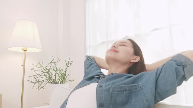 A relaxed young Asian woman sitting on a sofa, stretching her arms and resting with her eyes closed at home.