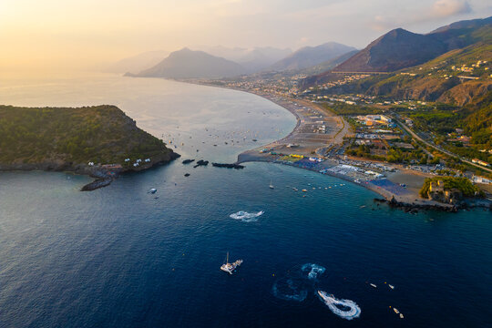 A breathtaking sunset aerial view captures Isola del Dino and the bay at Praia a Mare in Calabria, Italy. Boats navigate the deep blue waters along a sprawling beach, framed by dramatic coastal