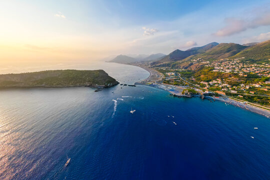 A breathtaking sunset aerial view captures Isola del Dino and the bay at Praia a Mare in Calabria, Italy. Boats navigate the deep blue waters along a sprawling beach, framed by dramatic coastal