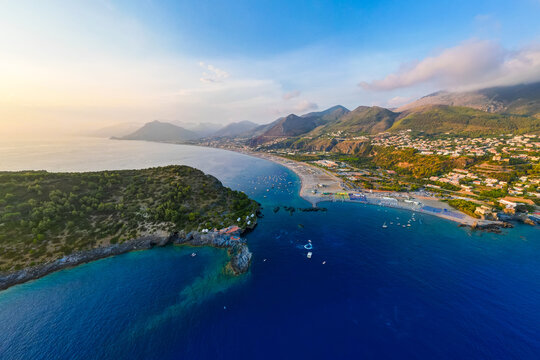 A breathtaking sunset aerial view captures Isola del Dino and the bay at Praia a Mare in Calabria, Italy. Boats navigate the deep blue waters along a sprawling beach, framed by dramatic coastal