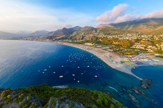 A breathtaking sunset aerial view captures Isola del Dino and the bay at Praia a Mare in Calabria, Italy. Boats navigate the deep blue waters along a sprawling beach, framed by dramatic coastal