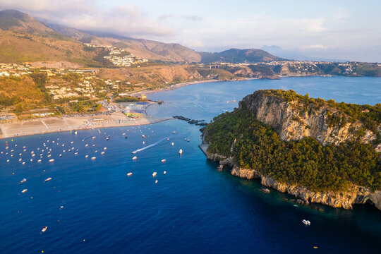 A breathtaking sunset aerial view captures Isola del Dino and the bay at Praia a Mare in Calabria, Italy. Boats navigate the deep blue waters along a sprawling beach, framed by dramatic coastal