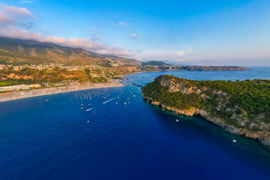 A breathtaking sunset aerial view captures Isola del Dino and the bay at Praia a Mare in Calabria, Italy. Boats navigate the deep blue waters along a sprawling beach, framed by dramatic coastal