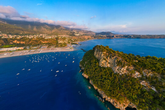 A breathtaking sunset aerial view captures Isola del Dino and the bay at Praia a Mare in Calabria, Italy. Boats navigate the deep blue waters along a sprawling beach, framed by dramatic coastal