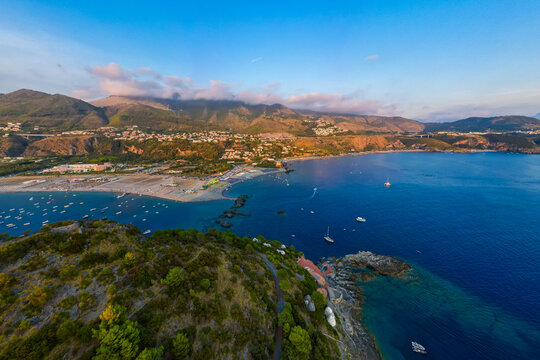 A breathtaking sunset aerial view captures Isola del Dino and the bay at Praia a Mare in Calabria, Italy. Boats navigate the deep blue waters along a sprawling beach, framed by dramatic coastal