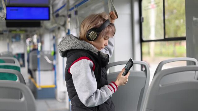 A teen girl rides public transport with headphones on, tapping and scrolling on her smartphone. Relatable urban commute scene illustrating digital habits, entertainment and everyday mobility.