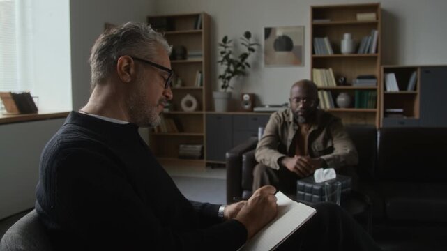 Middle aged Caucasian psychologist taking notes during therapy session with African American patient. Useful for mental health awareness campaigns and psychological counseling services