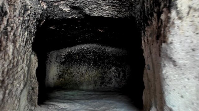 From a first-person perspective passing through a narrow, dark passage into an ancient burial chamber carved out of the rock, revealing the mysterious, crudely crafted interior of the historic catacom