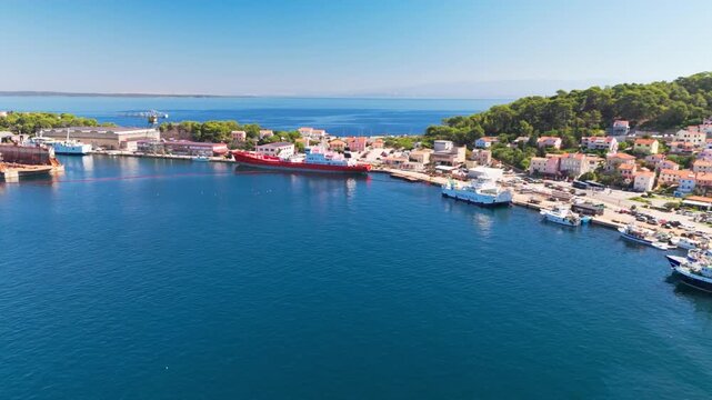 Drone turns above a bridge connecting the islands, revealing large ships, small boats and Mali Lo&scaron;inj spreading across the hills around a sparkling blue bay as a boat leaves a wake.