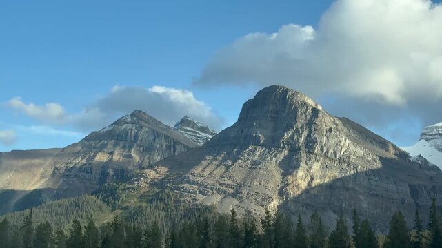 A serene view of the Rocky Mountains under a vast, clear blue sky, with patches of scattered clouds adding depth to the scene. The dense forest at the base of the mountains provides a lush foreground,