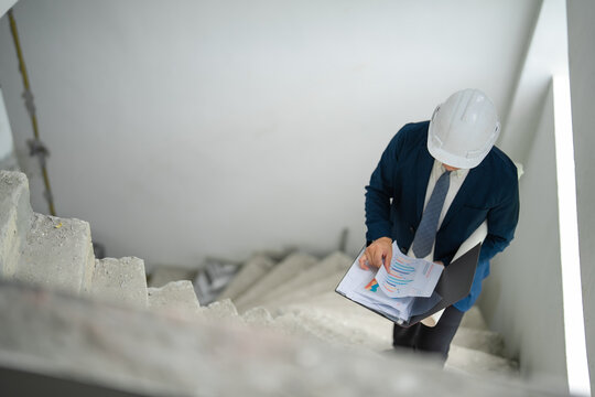 Construction manager reviews blueprint while walking on unfinished staircase, checking structural details, building progress and technical plans during site inspection.