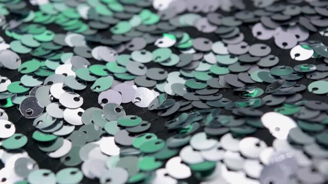 Close-up rotating shot of shimmering silver and green sequins on black fabric background, highlighting texture and light reflection