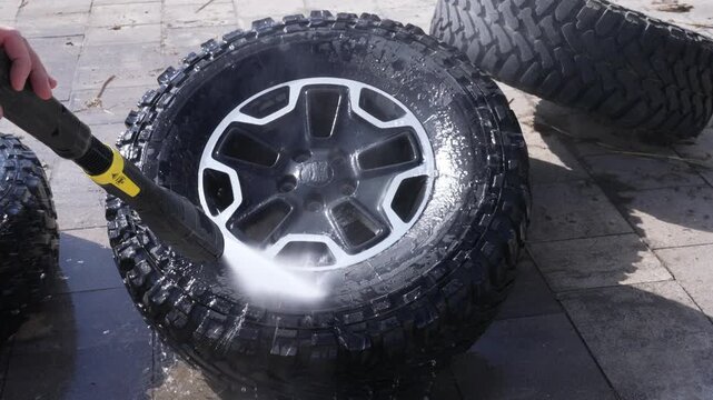Water jet cleans muddy tire tread. Black alloy rim gleams as spray hits. Tires sit near wet pavement. Clean wheels show rugged tread design. Ideal for vehicle maintenance scenes