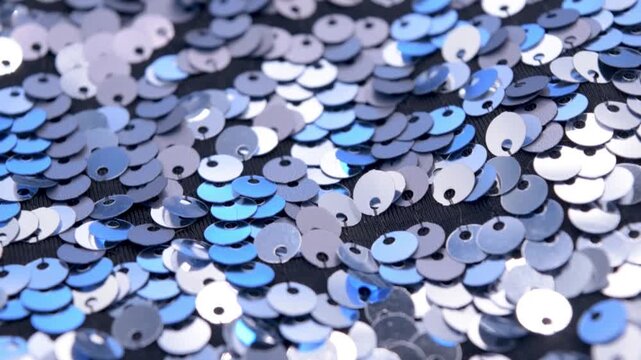 Close-up rotating shot of shimmering silver and azure sequins on black fabric background, highlighting texture and light reflection