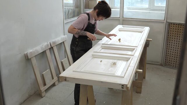 Attentive woman restorer removing wood putty paste excesses with chisel from vintage wooden door, focused female woodworker using carpentry tools and supplies in workshop, craftsmanship and handwork.
