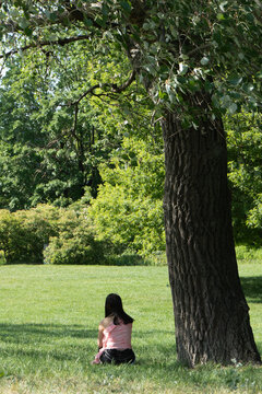 Lonely woman sitting on green grass under large tree in summer city park. Young female relaxing alone in public garden shadow. Mental health, wellness retreat and outdoor leisure concept.