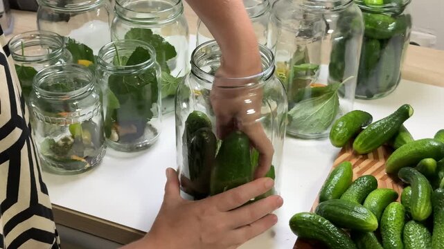a person handles a glass jar and places green cucumbers inside, surrounded by other filled jars and a pile of cucumbers on a wooden board